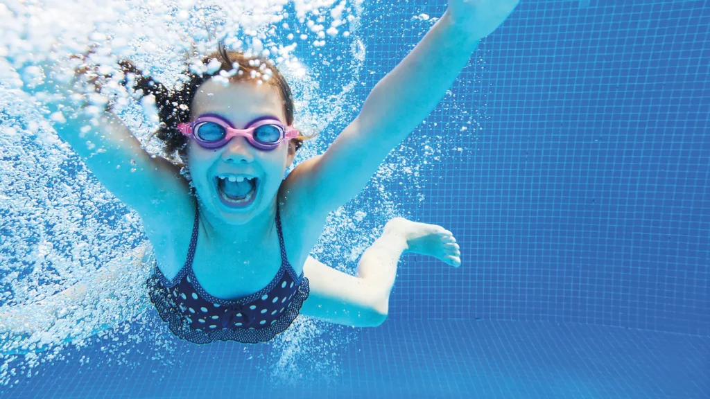 Ein Mädchen mit Schwimmbrille lacht unter Wasser in die Kamera